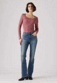 Γυναίκα που στέκεται μπροστά σε μονόχρωμο φόντο, φοράει ροζ παστέλ μακρυμάνικη μπλούζα, ψηλόμεσο μπλε τζιν και μαύρα μπότες με τακούνι.