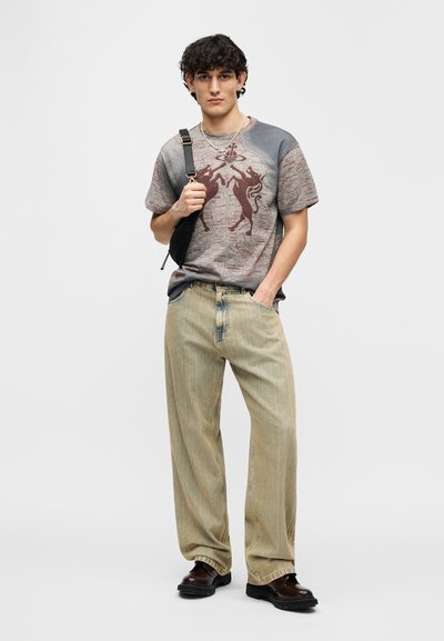 Γκρι και μπορντό γραφικό μπλουζάκι με αφηρημένο σχέδιο, συνδυασμένο με ανοιχτόχρωμο ριγέ παντελόνι με φαρδιά μπατζάκια και μαύρα παπούτσια· το μοντέλο κρατά μια μικρή τσάντα.