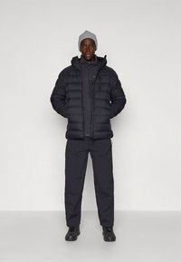 Juoda pūkinė striukė su užtrauktuku priekyje, dėvima ant tamsiai pilkos vidinės sluoksnio, derinama su juodomis krovinių kelnėmis ir pilka kepure.