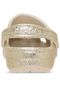 Błyszczące złote klapki o teksturowanym wykończeniu i szerokim pasku. Podeszwa jest biała i ma małe otwory wentylacyjne.