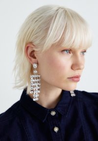 Orecchini a chandelier in argento con numerosi gemme trasparenti disposte in un motivo verticale. Modella con capelli corti biondo chiaro, che indossa una camicia scura.