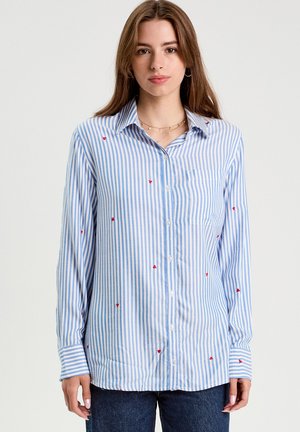 Ung kvinde iført en lyseblå og hvid vertikalt stribet skjorte med små røde hjerteprint og mørke jeans.