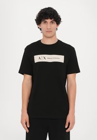 Svart bomull T-skjorte med korte ermer, med et kremfarget rektangulært logopanelsom viser "A/X ARMANI EXCHANGE" i svart skrift.