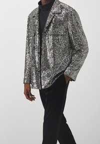 Blazer in paillettes argento con un fit rilassato, rever a contrasto e due tasche frontali, abbinato a un dolcevita nero e pantaloni neri.