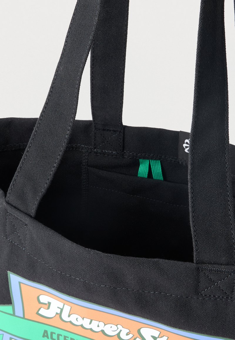 Bolsa tote de tela negra con asas reforzadas y una etiqueta impresa y colorida que dice "Flower Store." Bucle de tela verde dentro del bolsillo.