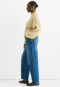 Cream cable-knit sweater, high neck, oversized fit; blue wide-leg jeans; black flats; model standing sideways against a white background.