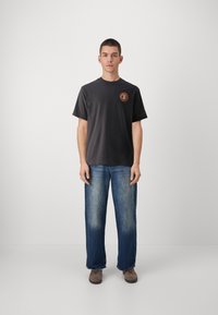 Dark gray cotton t-shirt with a circular orange logo on the chest, paired with blue denim jeans and brown shoes. Plain background.