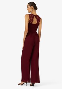 Burgunderfarbener Jumpsuit mit Spitze an den Schultern, offenem Rücken, gerafftem Taillenbund und weit geschnittenen Beinen. Glatte Textur und fließender Stoff.