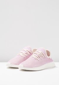 Pár světle fialových síťovinových tenisek s bílými podrážkami a šňůrkami, vystavených na bílém povrchu proti jednoduchému šedému pozadí.