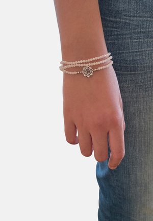 Mano appoggiata accanto alla gamba con jeans blu, adornata da un braccialetto a tre fili di perline bianche con un ciondolo rotondo in argento.