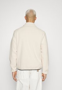 Fleece-pullover i ljus kräm med hög krage, avslappnad passform och svart kant vid nederkanten. Märke syns i nedre vänstra hörnet.