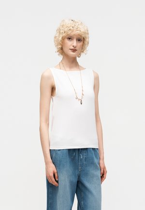 Ξανθιά γυναίκα με κατσαρά μαλλιά, που φοράει ένα λευκό αμάνικο τοπ, φαρδιά μπλε τζιν και ένα μακρύ κολιέ με μικρά φυλαχτά.