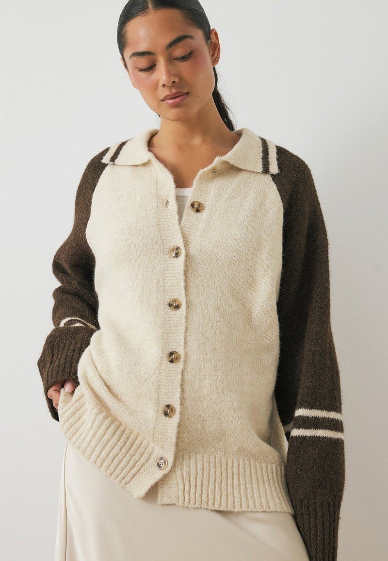 Pogu augšpusē atvērts kardigans ar krēma un brūna vilnas kombināciju, ar ribotām malām. Brūnās piedurknes ir ar baltām svītrām. Tekstūrēta auduma sastāvs nodrošina siltumu.