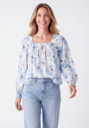 Kvindelig smilende iført lys blå blomsterlangærmet bluse med smocket halsudskæring og blå jeans mod en ensfarvet hvid baggrund.