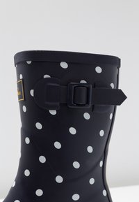 Musta kumisaapas, jossa on suuret valkoiset pilkut. Varressa on tekstuuria ja sivussa säädettävä musta solki.