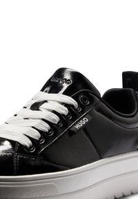 Schwarze Lackleder-Sneaker mit weißen Schnürsenkeln, gepolsterter Sohle und einem Logo an der Seite; mit Metallösen und einer strukturierten Oberfläche.