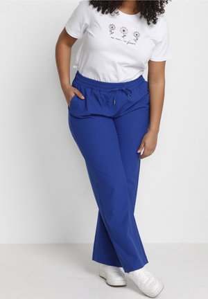 Frau trägt weißes T-Shirt mit drei lila Blumen und Text, blaue elastische Hose mit Kordelzug in der Taille und weiße Turnschuhe, steht an einer weißen Wand.