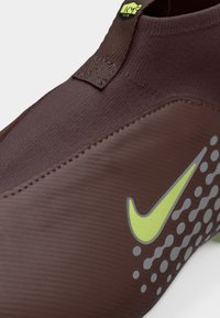 Makro posnetek rjave športne superge tipa slip-on z rumenim znakom Nike in odsevnimi pikami na teksturirani bočni plošči.