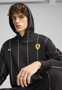 Crna dukserica s kapuljačom i bijelim vertikalnim prugama, s žutim logotipom Ferrarija prišivenim na prednjoj strani i malim izvezenim logotipom Pume na prsima.