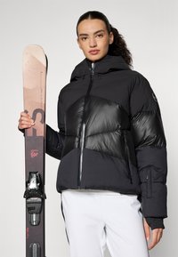 Schwarze Pufferjacke aus gemischten Materialien, mit glänzender Vorderseite und gerippten Bündchen. Hält einen braunen Ski mit schwarzen Bindungen und Markierungen.