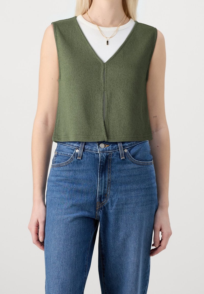 Bez rękawów zielony prążkowany crop top z dekoltem w kształcie litery V i rozcięciem z przodu, noszony na białej bluzce z długim rękawem. Połączony z niebieskimi jeansami o wysokim stanie.