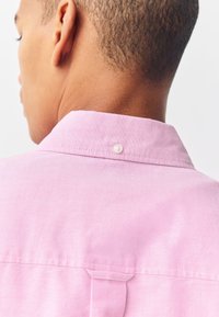 Camicia rosa con bottoni e collo a camicia, con spalla posteriore e chiusura con un solo bottone al collo. Tessuto dalla texture liscia.