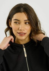 Sweat à capuche noir zippé avec une texture lisse, doté d'une fermeture éclair métallique et d'une étiquette. Le tissu semble doux et léger, avec un design minimaliste.