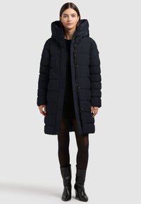 Mornarica puffer jakna s velikim, punjenim kapuljačom, prednjim patentnim zatvaračem i kopčanjem na dugmad. Tekstuirana tkanina s horizontalnim šavovima.