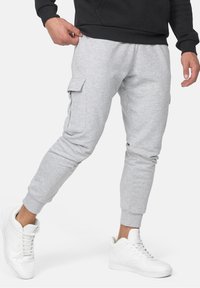 Grijze sweatpants met zijzakken, taps toelopende benen en elastische boorden, gecombineerd met witte sneakers op een neutrale achtergrond.