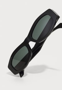 Gafas de sol negras con montura rectangular extragrande, lentes oscuros y un acabado mate suave. Presenta un diseño elegante y angular.