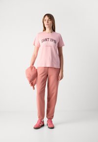 T-shirt di cotone rosa con stampa "GANT 1949 NEW HAVEN", abbinata a pantaloni sportivi rosa coordinati e sneakers rosa, mentre tiene una felpa rosa.