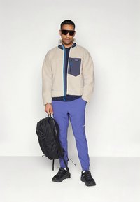 Casaco polar bege com detalhes azul-marinho, fecho azul e bolso no peito; combinado com calças roxas e mochila preta.