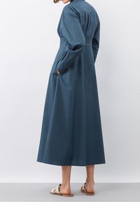 Un lungo vestito color turchese con colletto, maniche lunghe e tasche laterali, caratterizzato da una texture liscia e una vestibilità comoda in vita.