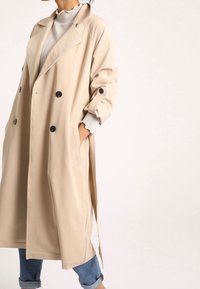 Beige vuorituuli takki laajalla kauluksella, kaksirivinen malli, suuret mustat napit ja sivutaskut. Vaatteena tekstiilistä valmistettu vaalea neule ja farkut.