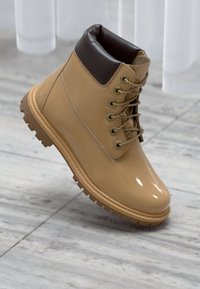 Stivale in pelle sintetica tan con un colletto imbottito marrone, occhielli in metallo dorato e suola in gomma strutturata. Presenta lacci robusti e un dettaglio del logo.