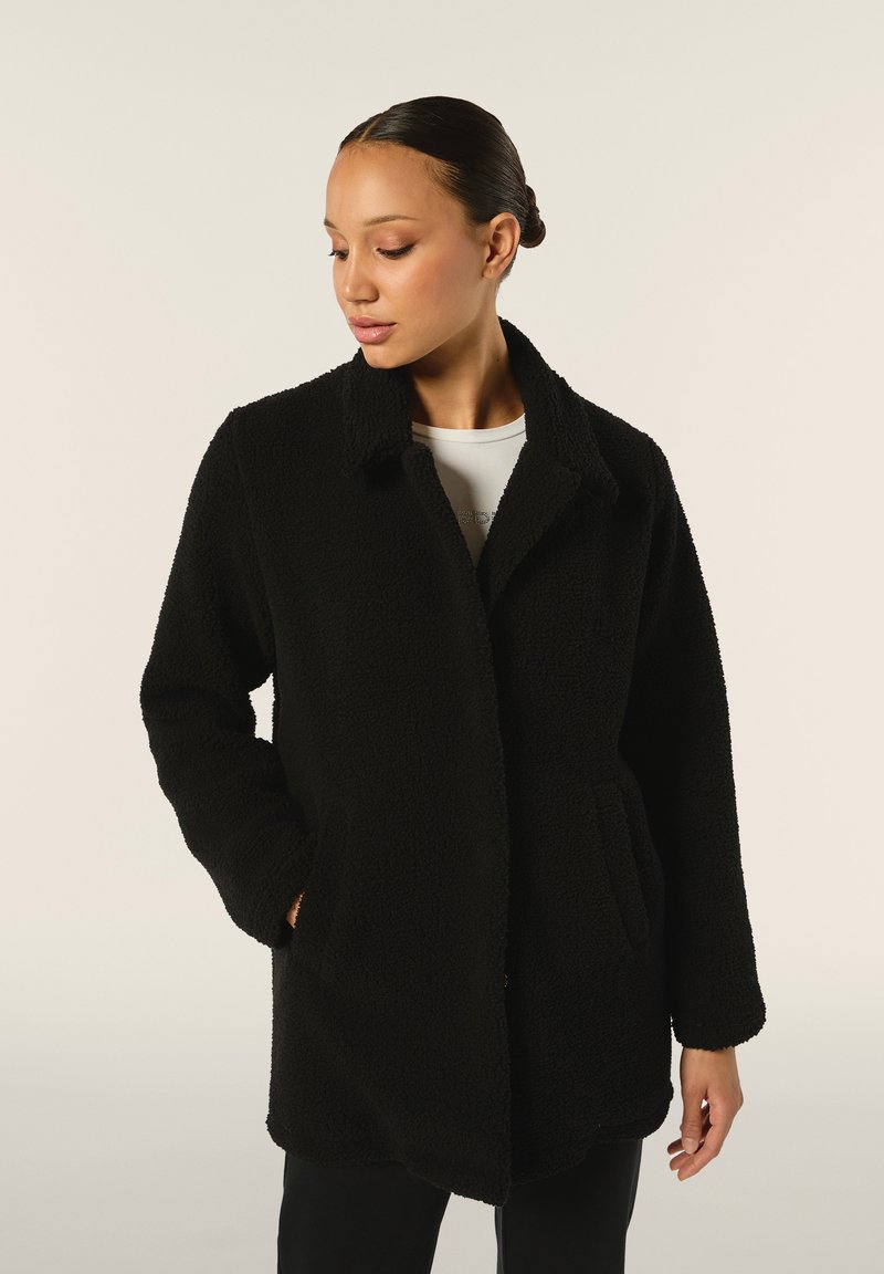 Cappotto nero peloso con colletto, chiusura a bottone e tasche frontali. Presenta un tessuto testurizzato e una vestibilità rilassata per il comfort.