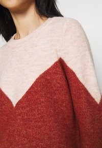 Stickad tröja i mjuk ullblandning med ett färgblock-design i ljusrosa och mörkröda nyanser, samt ett chevronmönster vid bröstet.