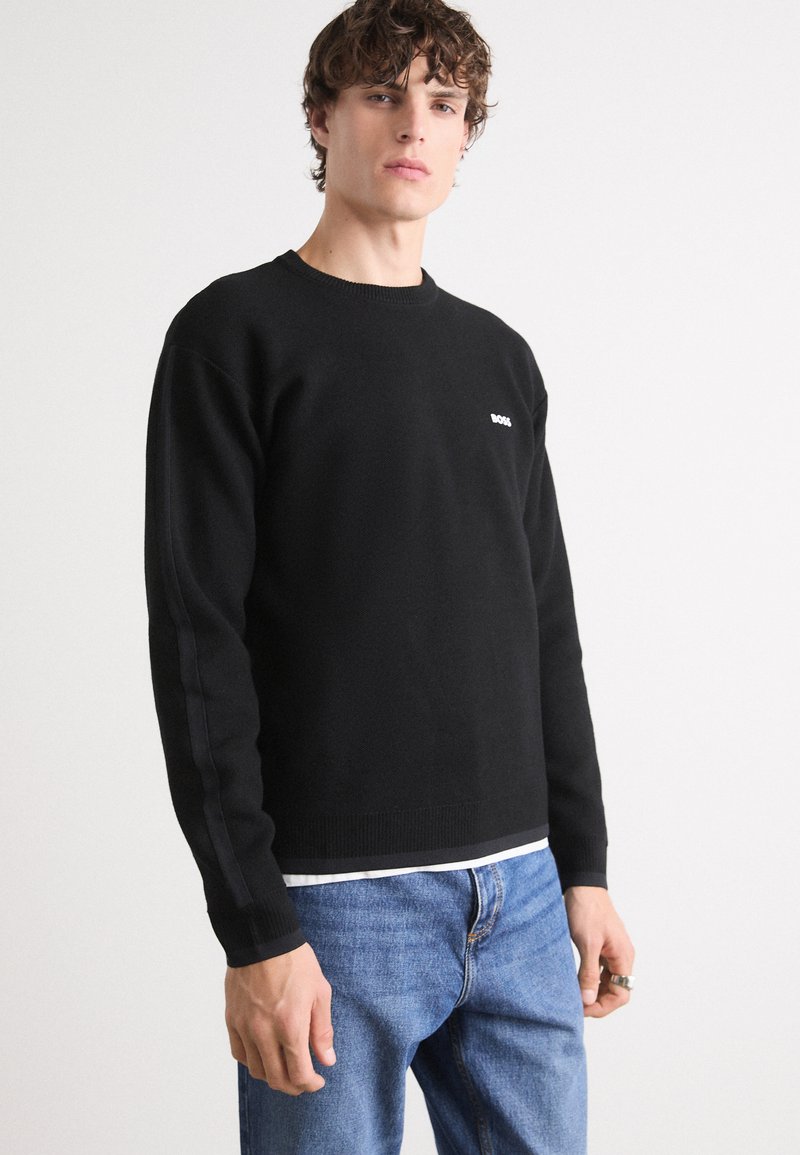 Czarny sweter dzianinowy z okrągłym dekoltem i ściśniętymi mankietami. Posiada małe białe logo na piersi. Noszony na białej koszulce i zestawiony z niebieskimi dżinsami.