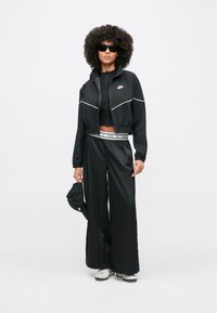 Μαύρο κοντό μπουφάν με λευκή διακόσμηση, ταιριαστόι μαύροι παντελόνες με φαρδιά πόδια και εφαρμοστό μαύρο τοπ. Αξεσουάρ περιλαμβάνουν μαύρα γυαλιά ηλίου και καπέλο.