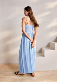 Robe longue bleue avec un corsage smocké, des bretelles fines et une jupe fluide plissée. Le mannequin se tient les mains jointes dans le dos.