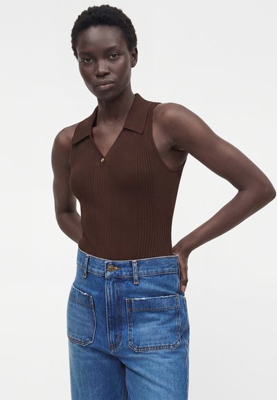 Γυναίκα με κοντά μαλλιά, που φοράει ένα καφέ μαν sleeveless ριπ τοπ και ψηλόμεσο μπλε τζιν με μεγάλες μπροστινές τσέπες, στέκεται με τα χέρια στη μέση.