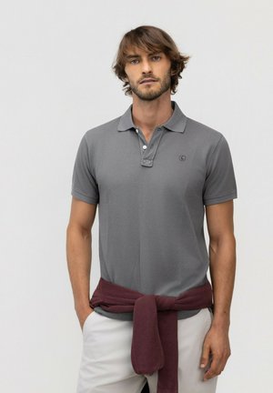 Hombre con cabello corto y castaño que lleva una camiseta polo gris y pantalones blancos con un suéter granate atado alrededor de su cintura, de pie frente a un fondo liso.