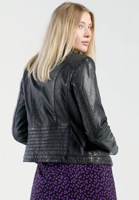 Zwarte leren jas met een getailleerd ontwerp, gestructureerde schouders, ribbelaccenten en horizontale stiksels op de rug. Draag een paarse met patroon rok eroverheen.