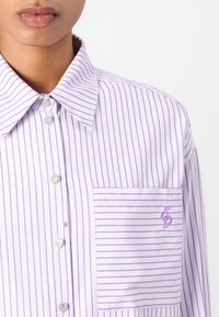 Femme portant une chemise blanche avec de fines rayures verticales violettes, des boutons en forme de cœur et une poche avant avec un logo brodé violet.