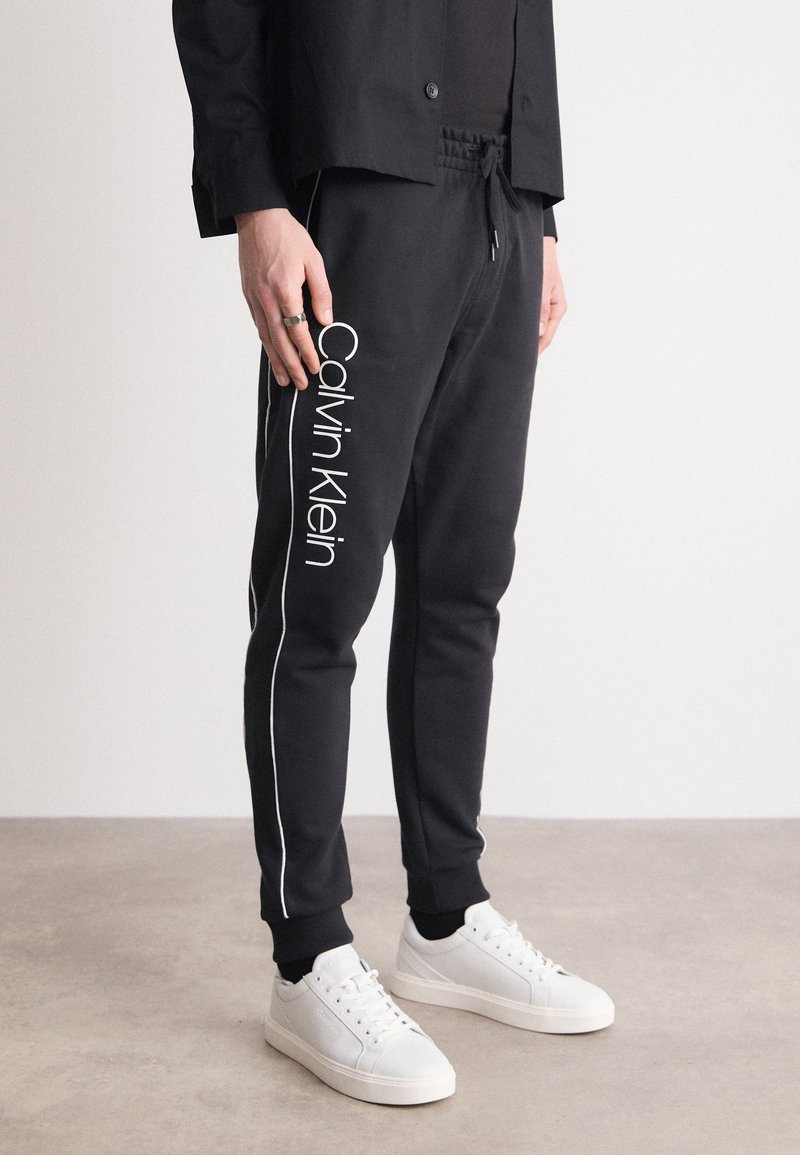 Black sweatpants with white side stripes and "Calvin Klein" logo on the left leg. Made from soft material, featuring an elastic waistband.