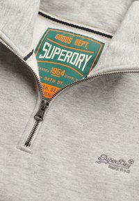 Jasnoszary sweatshirt na zamek o teksturowanym materiale. Posiada zieloną metkę z pomarańczowym i białym tekstem oraz srebrny zamek.