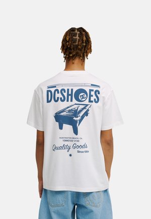 Osoba z krótkimi twistami w białej koszulce DC Shoes z niebieską grafiką stołu bilardowego i tekstem, w zestawie z jasnoniebieskimi dżinsami.