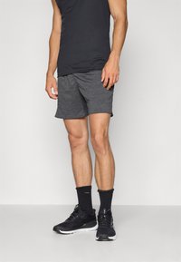 Calções de desporto cinzas com um cós elástico, combinados com sapatilhas pretas e meias até ao tornozelo pretas, sobre um fundo neutro.