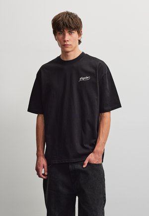 Jonge man met bruin haar, gekleed in een zwart oversized t-shirt en zwarte spijkerbroek, staand met één hand in zijn zak tegen een effen achtergrond.