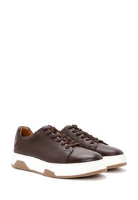 Un par de zapatillas bajas de cuero marrón oscuro con suelas gruesas blancas y suela marrón, dispuestas una al lado de la otra sobre un fondo blanco.